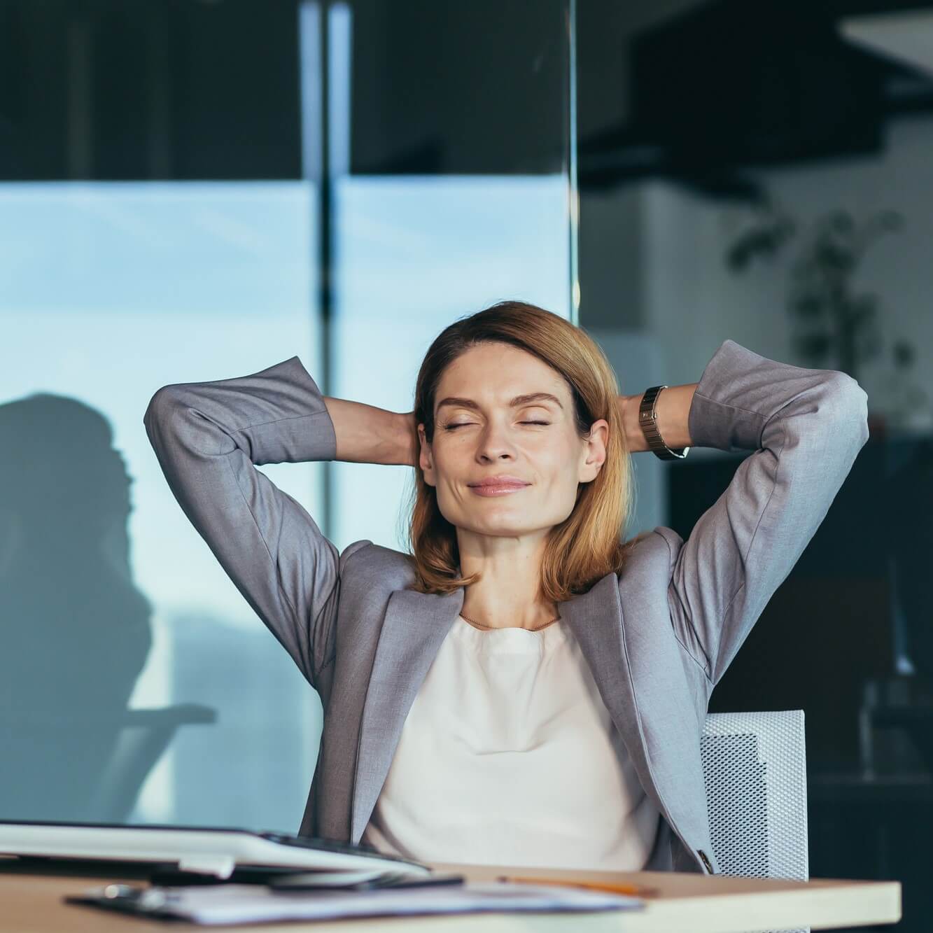 Entspannte Geschäftsfrau mit den Händen hinterm Kopf verschränkt, lächelnd mit geschlossenen Augen, genießt die Wirksamkeit der Atemtechniken.