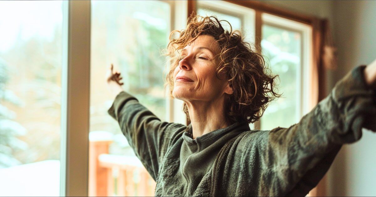 Eine Frau in den Wechseljahren steht am Fenster, breitet die Arme aus, atmet bewusst und tankt Tageslicht gegen Unruhe am Abend.