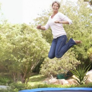Eine lachende Frau mittleren Alters springt auf einem Trampolin vor grünen Bäumen im Hintergrund, weil sie dank Beckenbode-Vital eine starke Mitte hat.